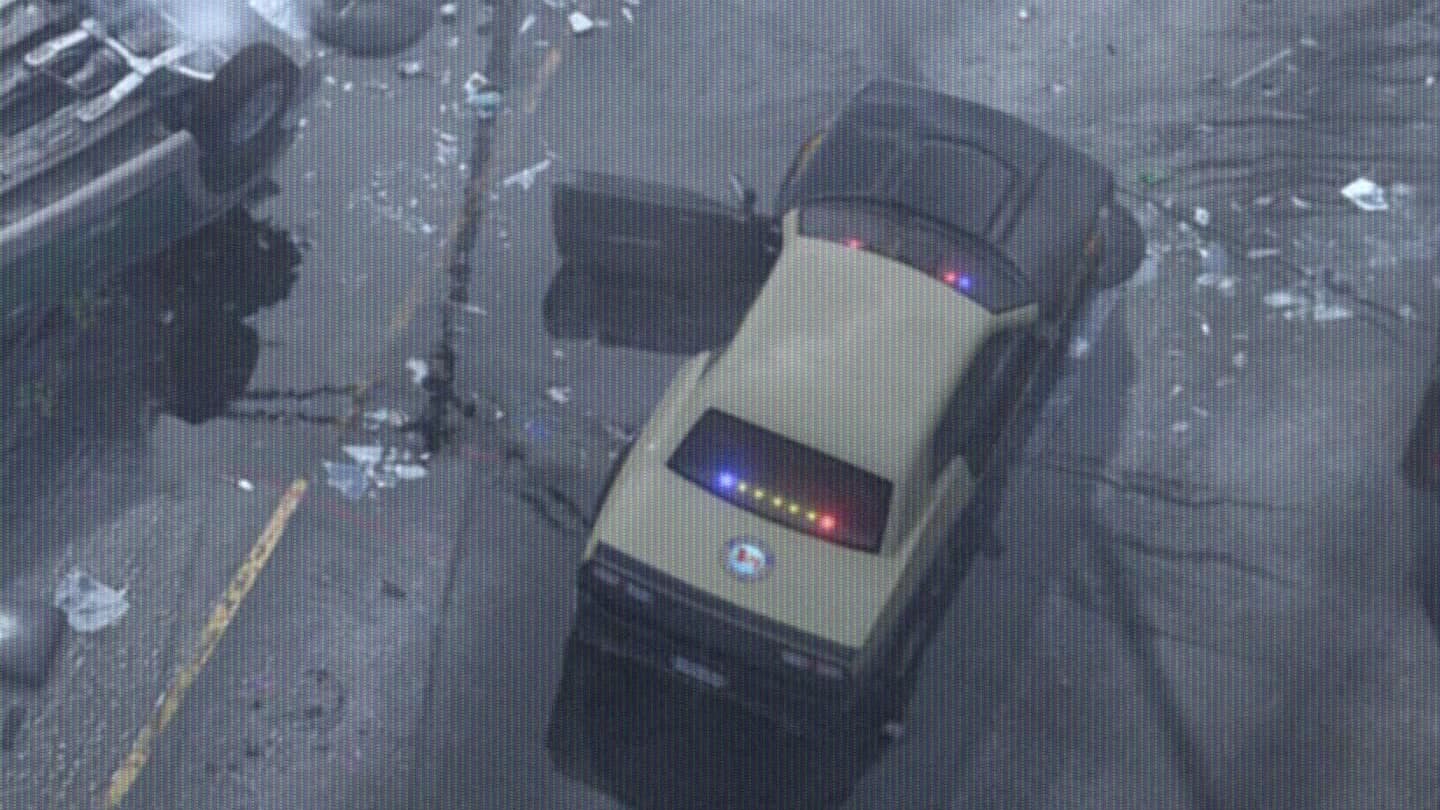 Beige muscle-car patrol unit with its lights on next to a highway crash, seen from a helicopter, in GTA VI Trailer 1