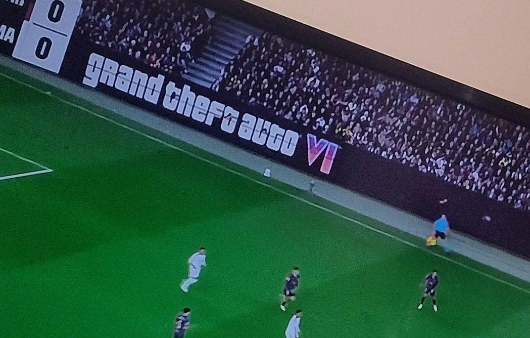 Photo of a TV screen: pitchside LED board with the 'grand theft auto VI' logo, packed stands behind, players and an assistant referee with a flag on the grass