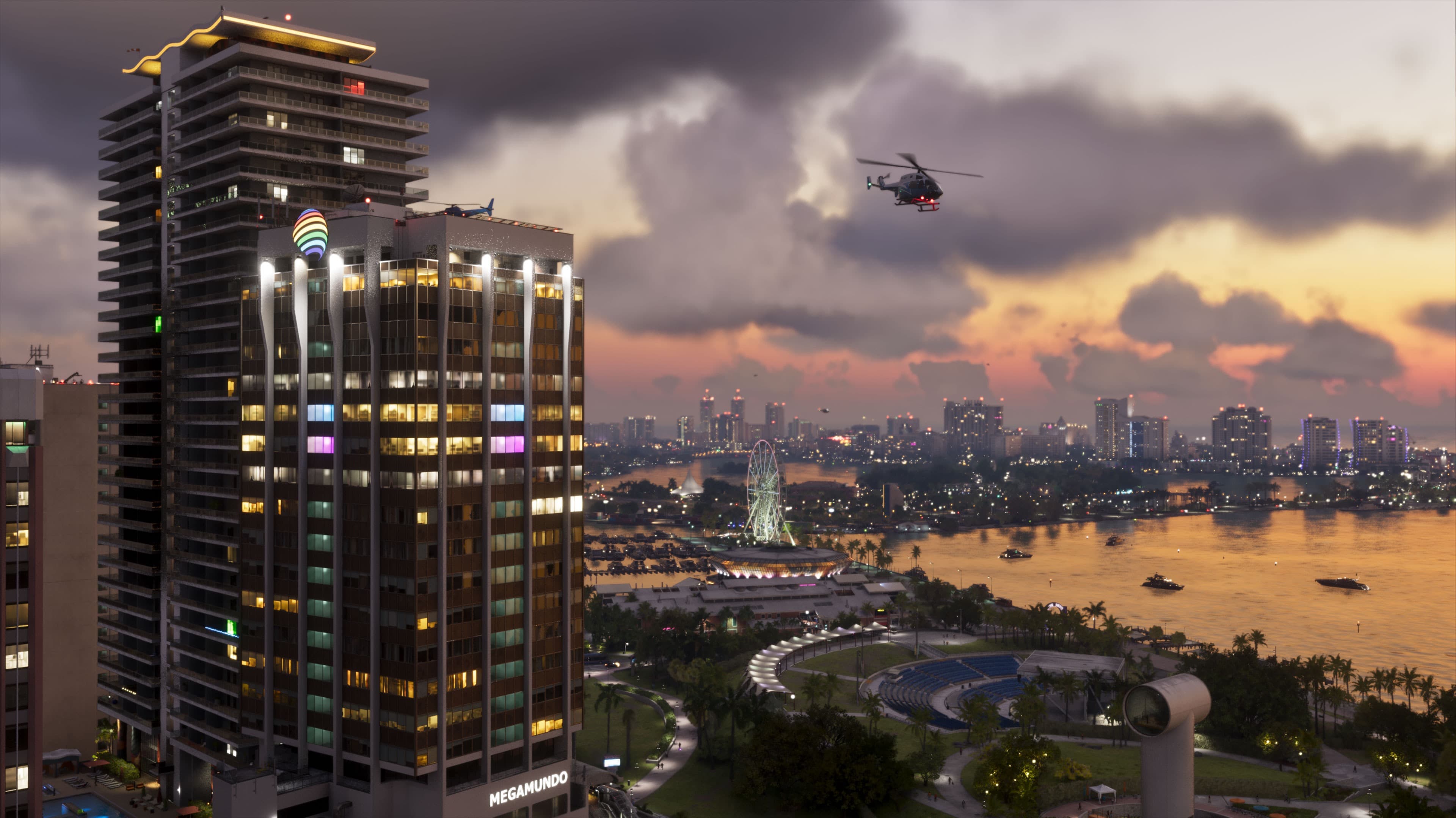 Atardecer tormentoso en Vice City: la torre iluminada Megamundo en primer plano, un helicóptero sobrevolándola y, al fondo, la bahía con la noria, barcos y el skyline.