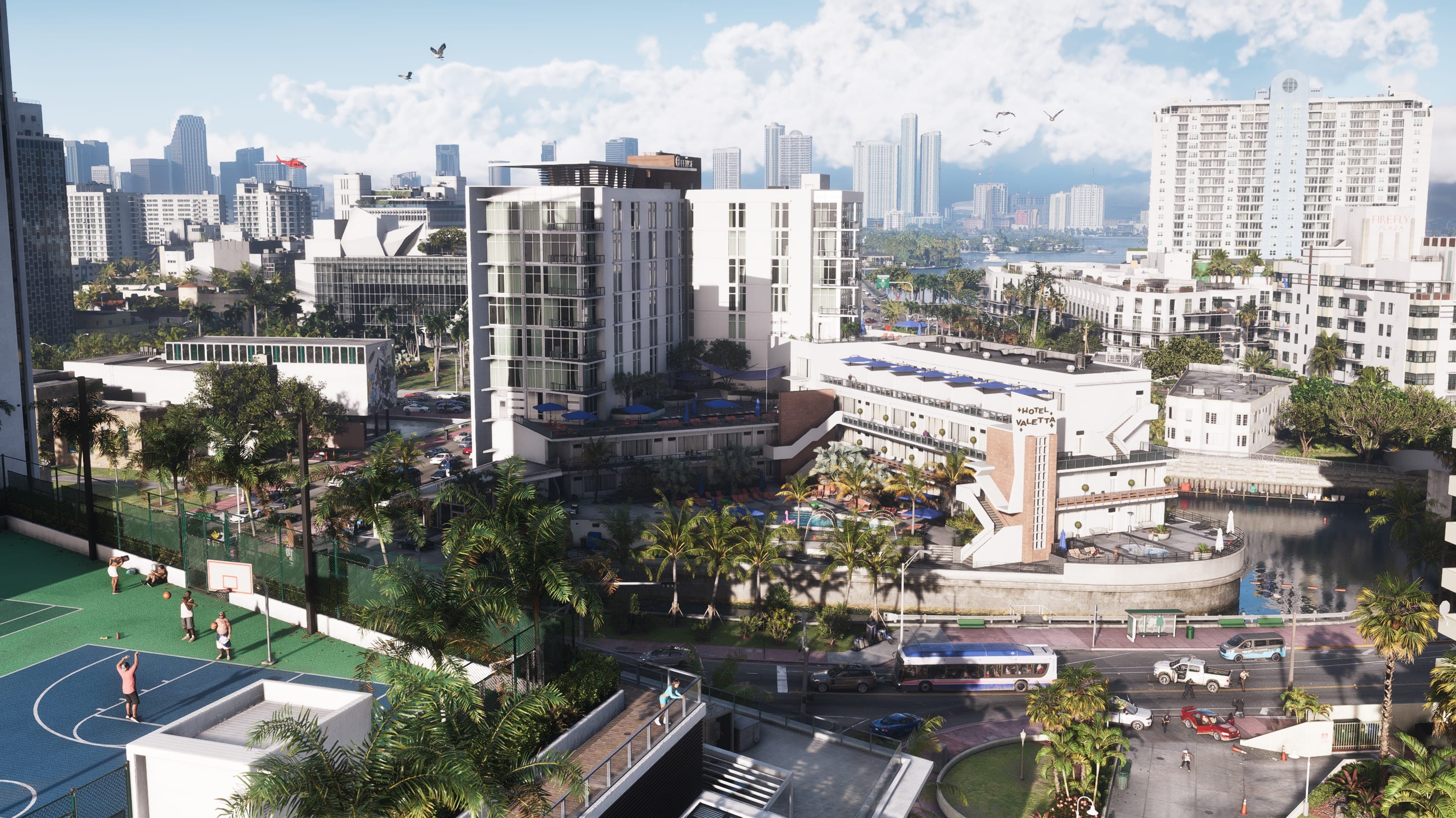 Vista aérea de día de Vice City en GTA VI: torres de apartamentos, una cancha de baloncesto, palmeras y tráfico, con el agua y los rascacielos del centro al fondo.